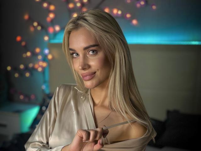 SabrinaSharm with blonde hair, smiling, wearing a silky beige top, pulling down the strap to reveal a bit of her tits. She has fair skin and cute, sexy eyes.