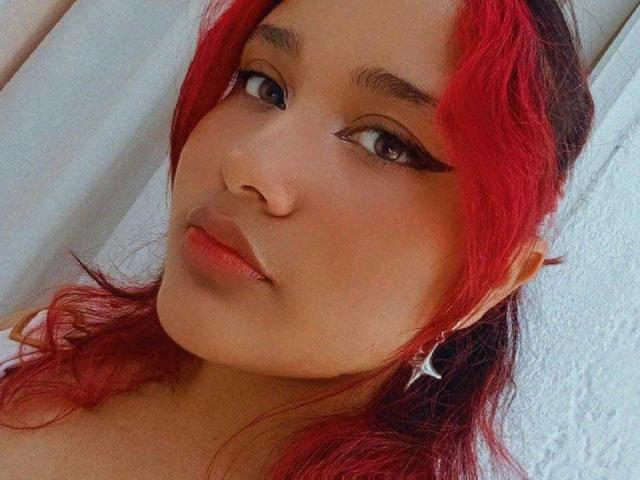 Nayra : Une femme aux cheveux roux, à la peau brune et avec un trait d'eye-liner noir, regardant vers le haut avec un léger sourire, portant un bandeau blanc et une boucle d'oreille en forme d'étoile argentée sur le côté droit..