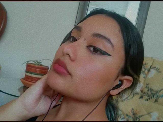 Nayra, met zwarte eyeliner, roze lippenstift en oordopjes, die haar kin aanraakt. Ze heeft een kleine piercing in haar gezicht en donker haar, en kijkt iets omhoog..