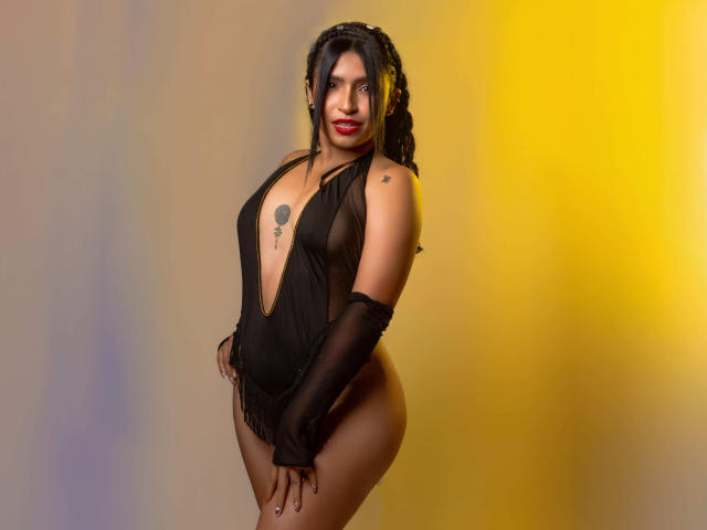 Conyngas with dark hair, red lipstick, and a deep black halter dress, showing her tits, ass, and a tattoo on her chest. She's wearing long black gloves.