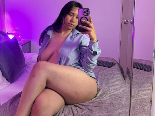 Karollteyes with big tits and curvy ass, wearing an open blue shirt, sitting on a bed, holding a phone, purple lighting, mirror reflection, taking a selfie.