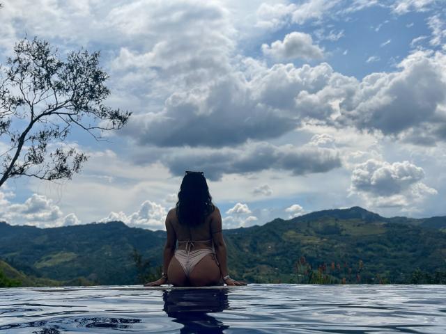 ElizabethMacy with dark curly hair, tan skin, and a bikini bottom, sitting in a pool, facing mountains and a cloudy sky, her ass slightly above water.