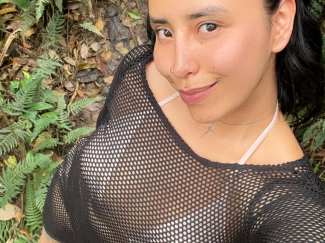 ElizabethMacy with tan skin, black mesh top, visible small breasts, nose piercing, smiling, brown eyes, black hair, ferns, and leaves in background, wearing a thin pink strap.