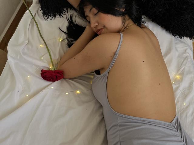 Elizaa is lying on a bed, back to the camera, wearing a gray dress, with a red rose and fairy lights. She has dark hair, fair skin, and a nose ring.