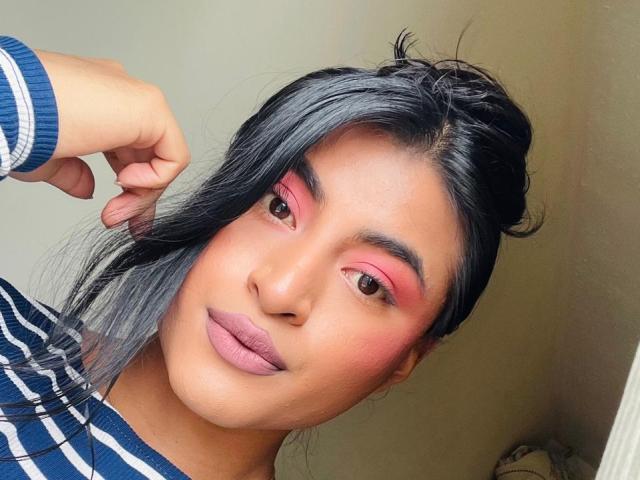 MichelleDenise with pink eyeshadow, gray-black hair, and striped shirt, touching her head with a hand, showing pink lips and a slight smile. Her ass is not visible.