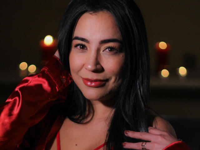 CyntiaSarat with black hair, red lipstick, and a red satin shirt, smiling softly, hand on neck, red candles in background, glowing softly. Her tits are small and cute.