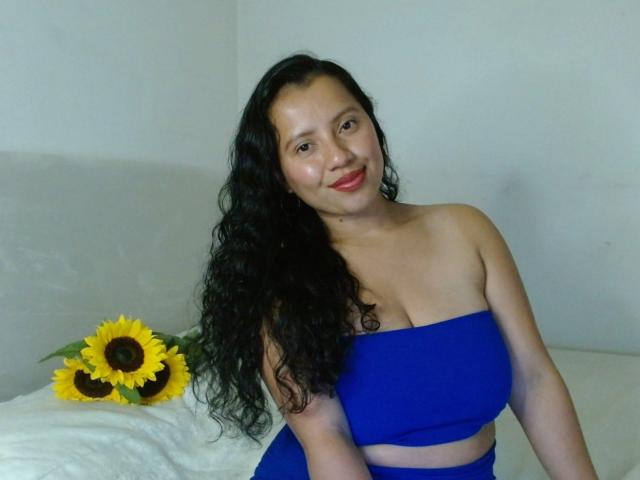 Isis69s with curly black hair, blue strapless top, and matching skirt, smiling, showing her tits, sitting with a sunflower on a white bed.