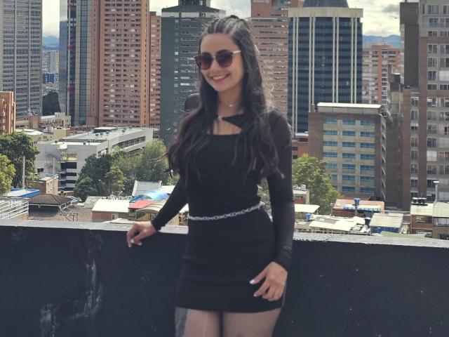 SophieJones69 stands on a balcony, wearing a tight black dress with long sleeves, a cutout neckline, and a silver belt, smiling with dark sunglasses, and wavy brown hair.