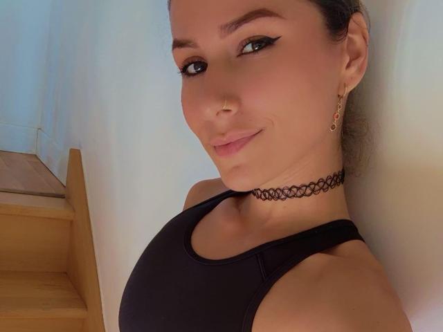 SophyaEvans with black eyeliner, nose ring, and choker, smiling at the camera, wearing a black tank top, showing her tits, and wooden stairs in the background.