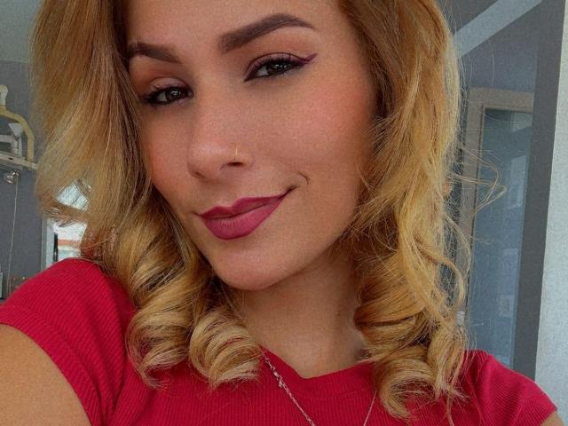 SophyaEvans has blonde curly hair, red lipstick, and winged eyeliner, wearing a red rib-knit shirt, with a small nose ring and a subtle smile. Her tits are not visible.