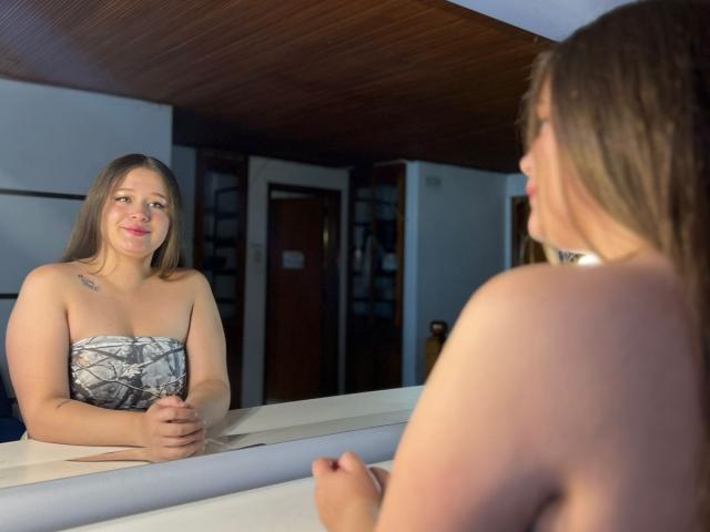 KellyHoty with small tits, smiling at her reflection in a mirror. She's wearing a strapless black and white patterned top, and has a tattoo on her left shoulder.