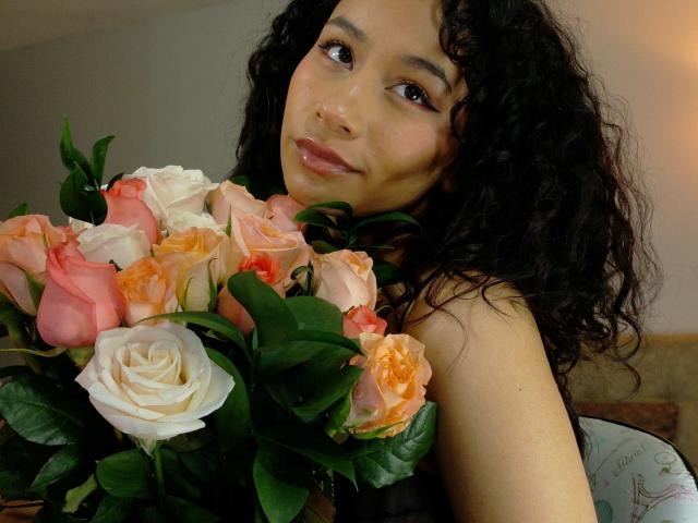 Heily Peterson, con cabello negro y rizado, sosteniendo un ramo de rosas rosadas y blancas, mirando a la cámara con una sonrisa sutil, con una luz natural suave..