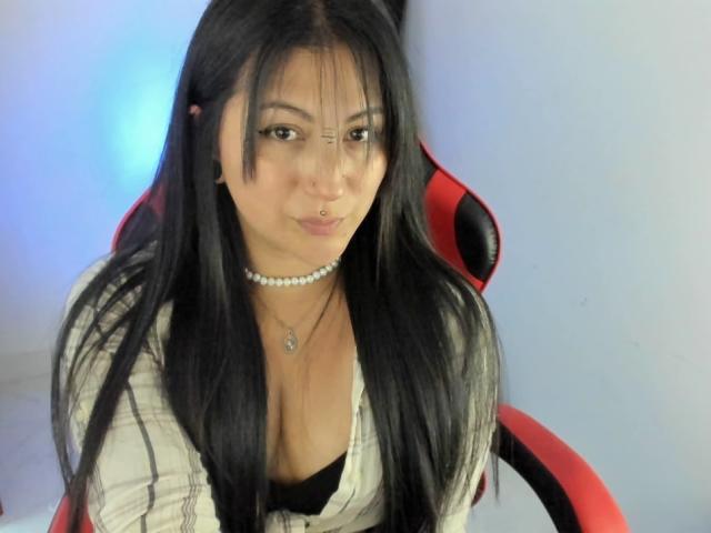 LilitthHot with long black hair, brown eyes, nose ring, pearl necklace, white plaid shirt, black bra, sitting in red and black chair, pouty expression, medium tits.