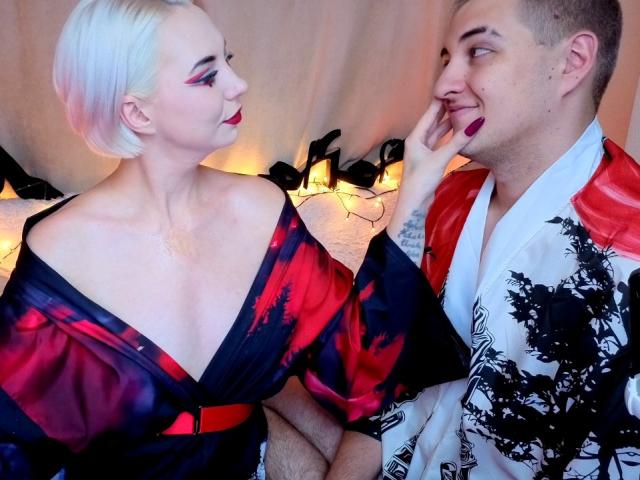 BlondIce with pale skin, short platinum blonde hair, and red lipstick, touches a man's face, wearing a red and black kimono, exposing her tits, he's in a white kimono.