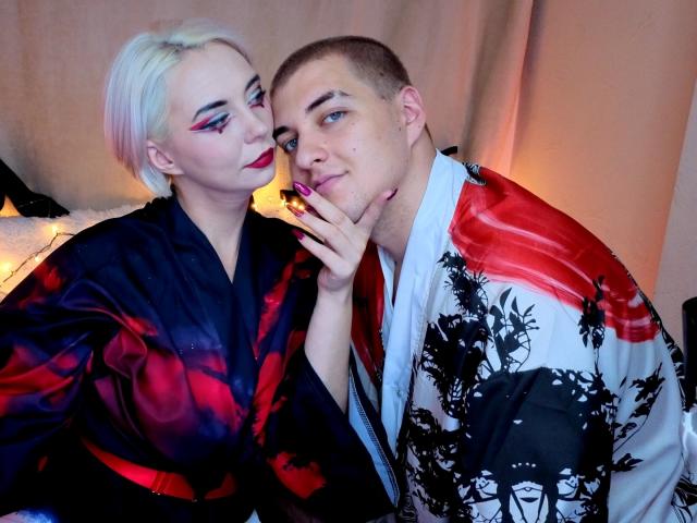 BlondIce with short blonde hair, wearing a black and red kimono, leans on a bald man in a white and red kimono, touching his cheek with her finger.
