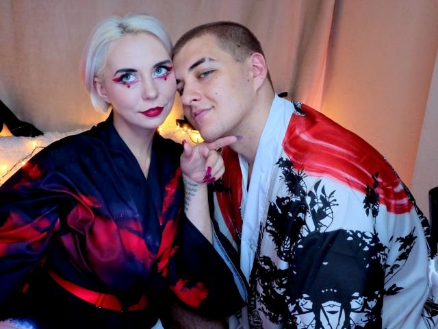 BlondIce with short platinum blonde hair and red lipstick, wearing a black-red floral robe, leans on a bald man in a white-red-black floral robe, pointing to his nose.