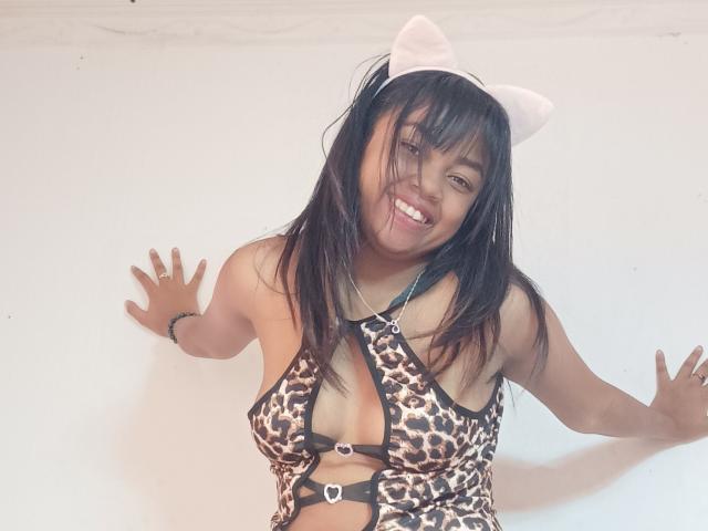 MaliciaQueen with cat ears, smiling, wearing a leopard-print halter top that exposes her small tits, against a white wall, hands on the wall, black hair, dark eyes.