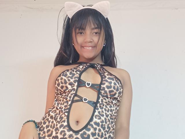 MaliciaQueen with dark hair, wearing a leopard print dress with cutouts, black straps, and white cat ears, smiling at the camera. Her tits are visible through the dress.