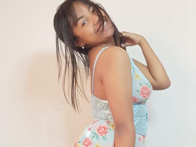 MaliciaQueen with dark straight hair, smiling, wearing a floral dress with white lace, showing her tits, standing sideways, hand near her head, white wall background.