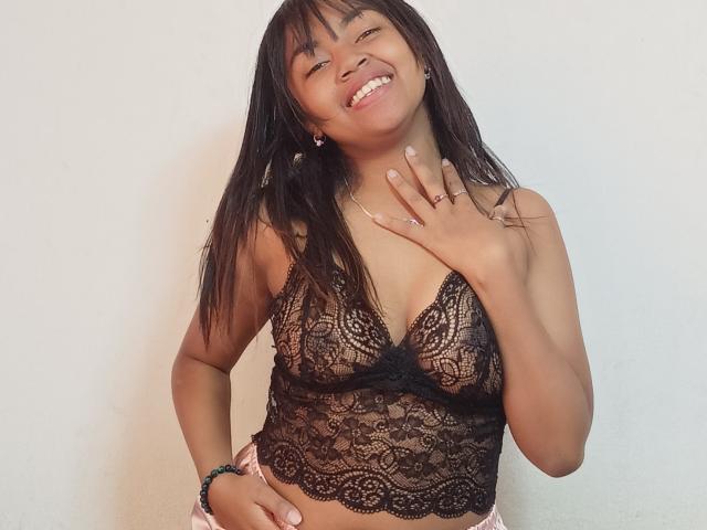 MaliciaQueen with dark lace bralette, showing her small tits, smiling, hand on chest, long black hair, pink bracelet, and gold ring.