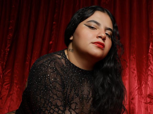 JannaFrank with long black wavy hair, red lipstick, black lace top, and subtle tattoo on neck, looking down with a slightly seductive expression. Red curtain backdrop.