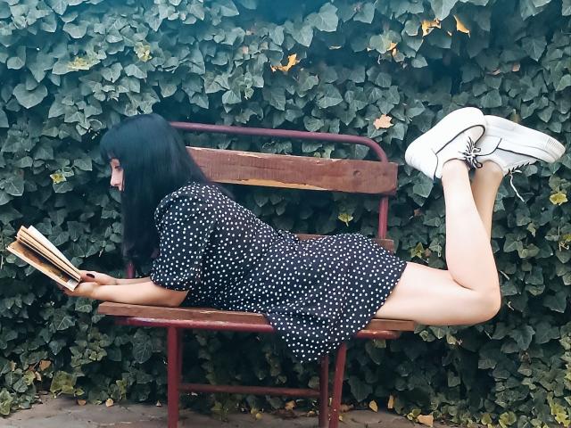 JillSilvies with black hair, lying on red bench, wearing polka dot dress, white sneakers, reading book, ass up, legs bent, against leafy green wall.