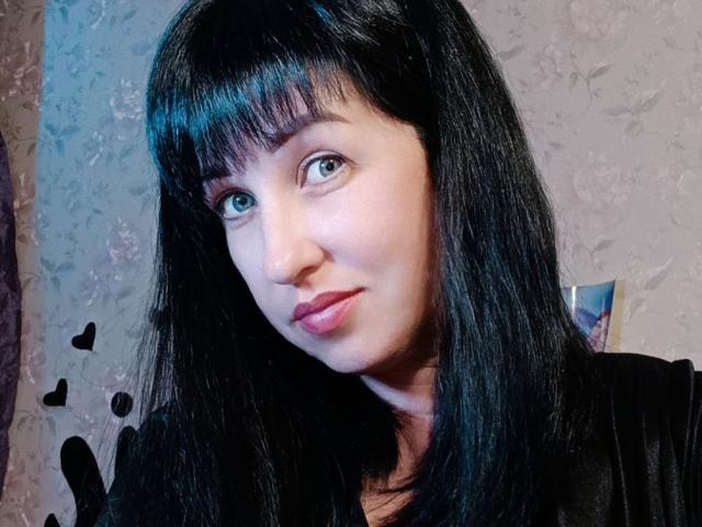 JillSilvies has black straight hair with bangs, pale skin, green eyes, and pink lips, wearing a black top, with small black heart stickers in the background.