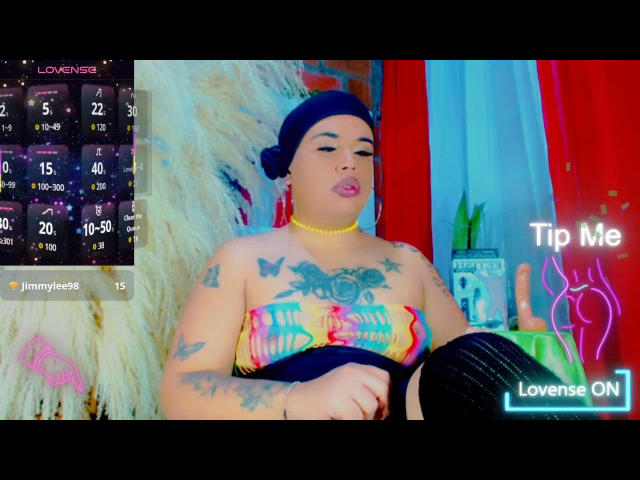 NauthyValentina with colorful strapless top, tattoos, yellow necklace, and blue headscarf, sitting with a relaxed expression, showing her tits and ass. Background includes a dildo on a shelf.