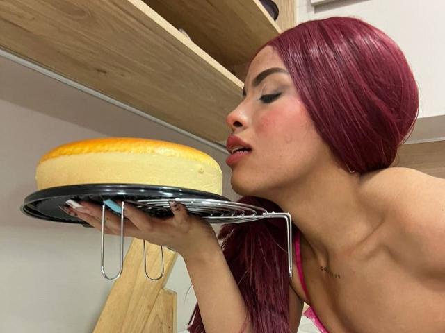 LexieBloom with purple hair, looking at a golden-brown cake on a metal shelf, holding it with her hand, wearing a pink strap, slightly open mouth, visible collarbone.