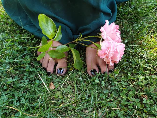 LadyNocturne's voeten, met zwarte nagellak, houden een roze roos met groene bladeren vast, op het gras. Haar groene broek is lichtelijk zichtbaar..