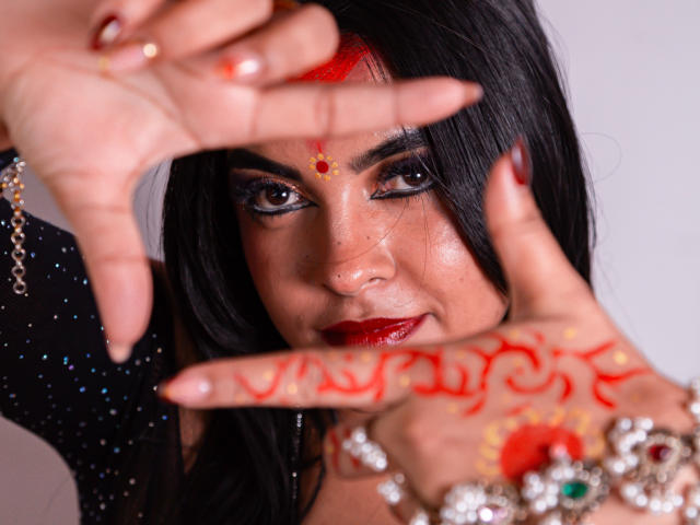 LakshmIrani's dark-haired face with red henna on her hands, wearing red lipstick, black glittery dress, and jewelry, holding her hands in front, showing intricate henna patterns.