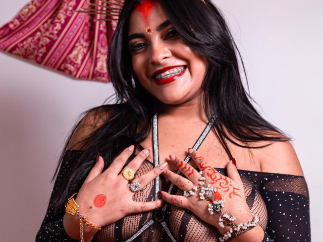 LakshmIrani with dark hair, red face paint, and red lipstick, smiling, holding her mesh-covered tits, wearing rings and jewelry, white background, red patterned cloth behind.