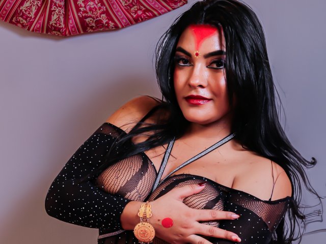 LakshmIrani with black hair, red forehead dot, and red lipstick, wearing a sheer black top, covering her tits with one hand, gold bracelet, and red nail polish, smiling.