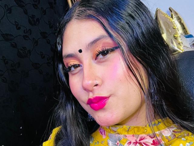 KalpanaAisha has black hair, pink lipstick, and eyeliner with a small black dot on her forehead. She's wearing a yellow floral top and has a nose ring.