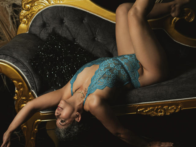 JuliaNewell is lying on a dark, velvet couch with gold trim, wearing a teal lace bodysuit, exposing her tits, with her legs up and arms stretched, in a sexy pose.
