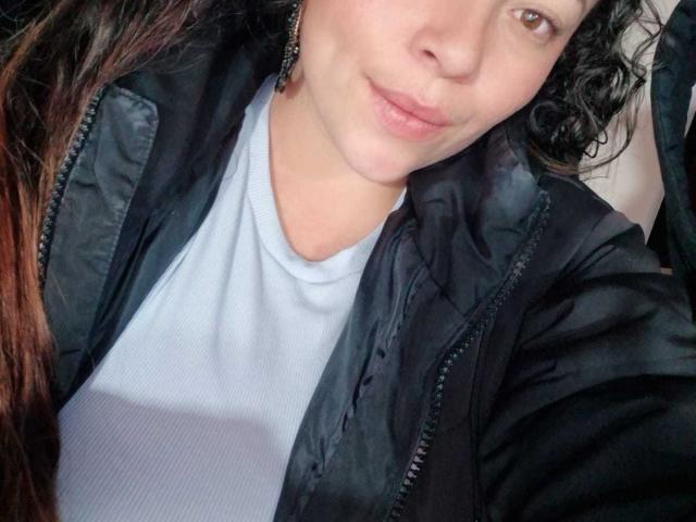 HoneyHotX with curly dark hair, light skin, and pouty lips, wearing a black jacket over a white ribbed shirt. Her brown eyes are slightly visible.