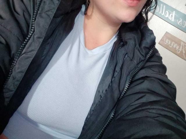 HoneyHotX indossa una giacca imbottita grigia sopra una camicia a coste azzurra, che lascia intravedere leggermente il seno, con capelli scuri ondulati e labbra rosa pallido..