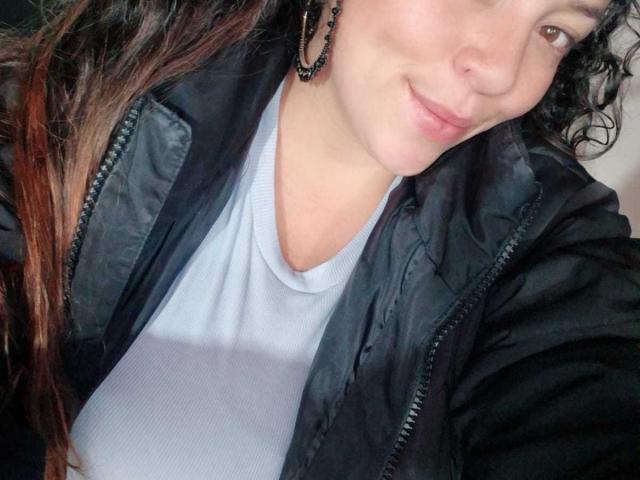 HoneyHotX with curly brown hair, wearing a black jacket over a white shirt, smiling slightly, showing a cute nose, and wearing black earrings in a selfie.