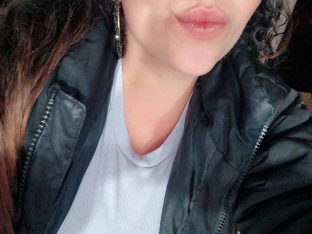 HoneyHotX mit sinnlichen, rosafarbenen Lippen, einer schwarzen Lederjacke, einem weißen T-Shirt und lockigem, braunem Haar, trägt auffällige Ohrringe. Ihre zarte, helle Haut steht im Kontrast zu ihrer dunklen Jacke..