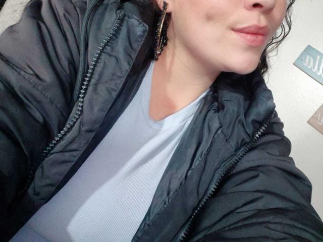 HoneyHotX mit voller Lippe, trägt eine schwarze, gepolsterte Jacke über einem weißen, gerippten Hemd, schwarze Ohrringe und leicht gewelltes, dunkles Haar, Gesicht teilweise sichtbar..