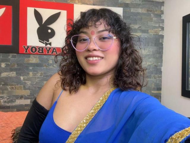 ShailaYojana with curly black hair, pink glasses, and blue dress with gold trim, smiling, showing her boobs, in a room with stone wall. She's wearing black gloves.
