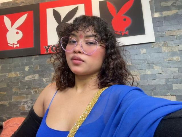 ShailaYojana with curly dark hair, pink glasses, and blue sheer top with gold trim, showing off her small tits, sitting in a room with stone walls.