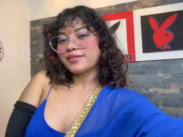 ShailaYojana with curly black hair, pink glasses, red dot on forehead, blue dress with gold trim, black sleeve, smiling, medium tits, cute, dark stone wall background.