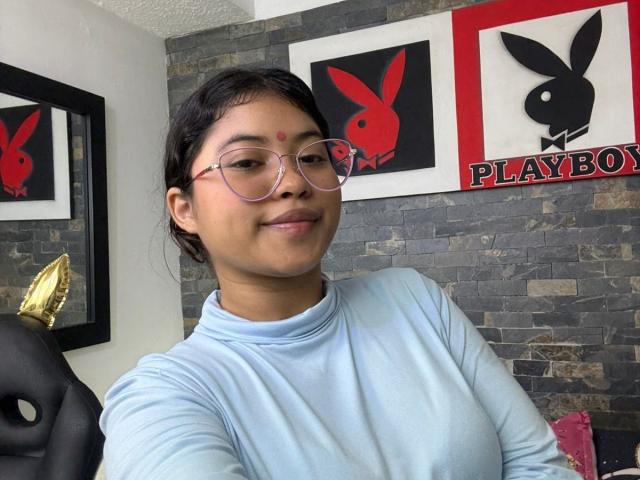 ShailaYojana with glasses, light blue shirt, and a red bindi, smiling at the camera, showing her small, cute ass, in a room with Playboy posters.