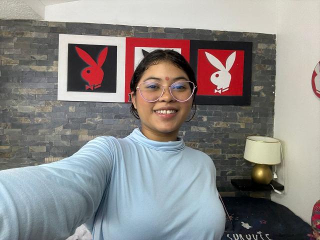ShailaYojana with short dark hair, glasses, light blue turtleneck, smiling, showing her tits, standing in front of three red and black Playboy posters, stone wall background.