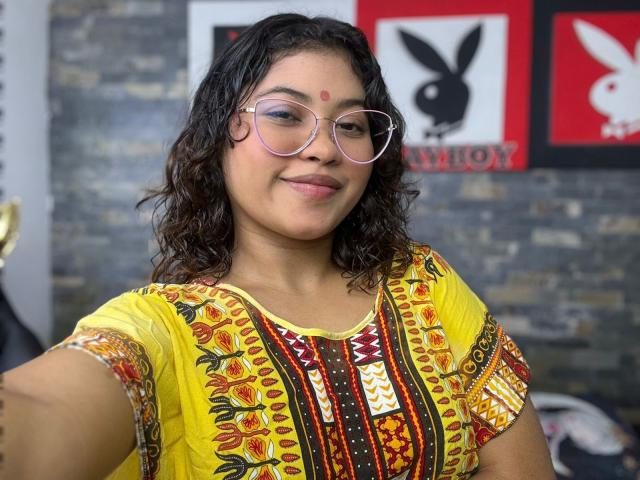 ShailaYojana with curly brown hair, pink glasses, yellow embroidered top, red dot on forehead, smiling, medium tits, cute, dark skin, standing in front of a wall with Playboy logos.