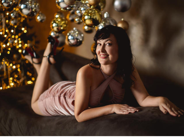 NathalyStars is laying on a couch, wearing a pink, criss-cross dress, black heels, with her tits slightly visible, smiling, and her ass raised, surrounded by shiny ornaments.