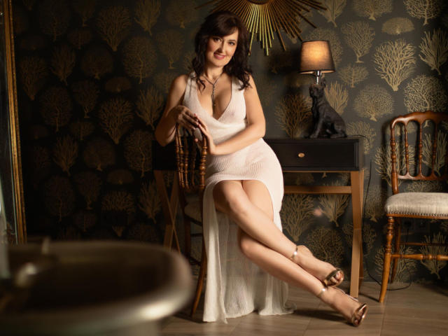 NathalyStars, with her tits in a white, deep-cut dress, sits on a chair, legs crossed, smiling, wearing sandals, in a dark room with a sunburst clock.