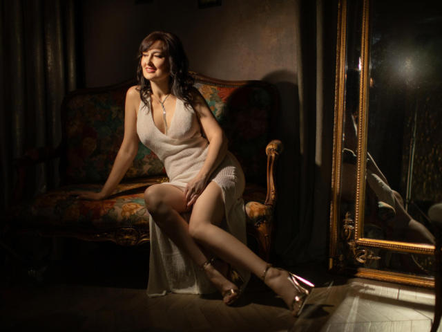 NathalyStars with wavy brown hair, wearing a sheer white dress, sitting on a floral couch, showing her tits, with pink heels, and a reflection in a gold mirror.