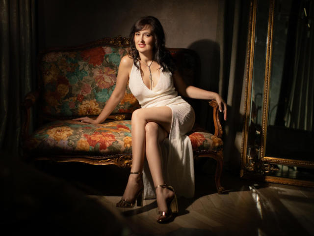 NathalyStars with medium tits, wearing a white dress, sitting on a floral couch, legs crossed, gold heels, dark curly hair, smiling, dimly lit, elegant, sexy, ornate mirror.